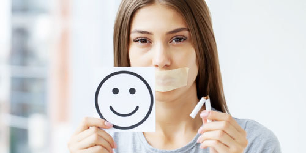 Chica con la boca tapada enseñando un cigarrillo partido por la mitad y una carita feliz dibujada en un papel